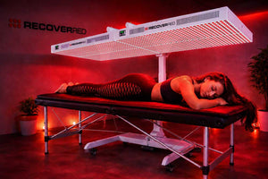 Person lying on a massage table under a large LED light with 'RecoverED' branding in a dimly lit room.